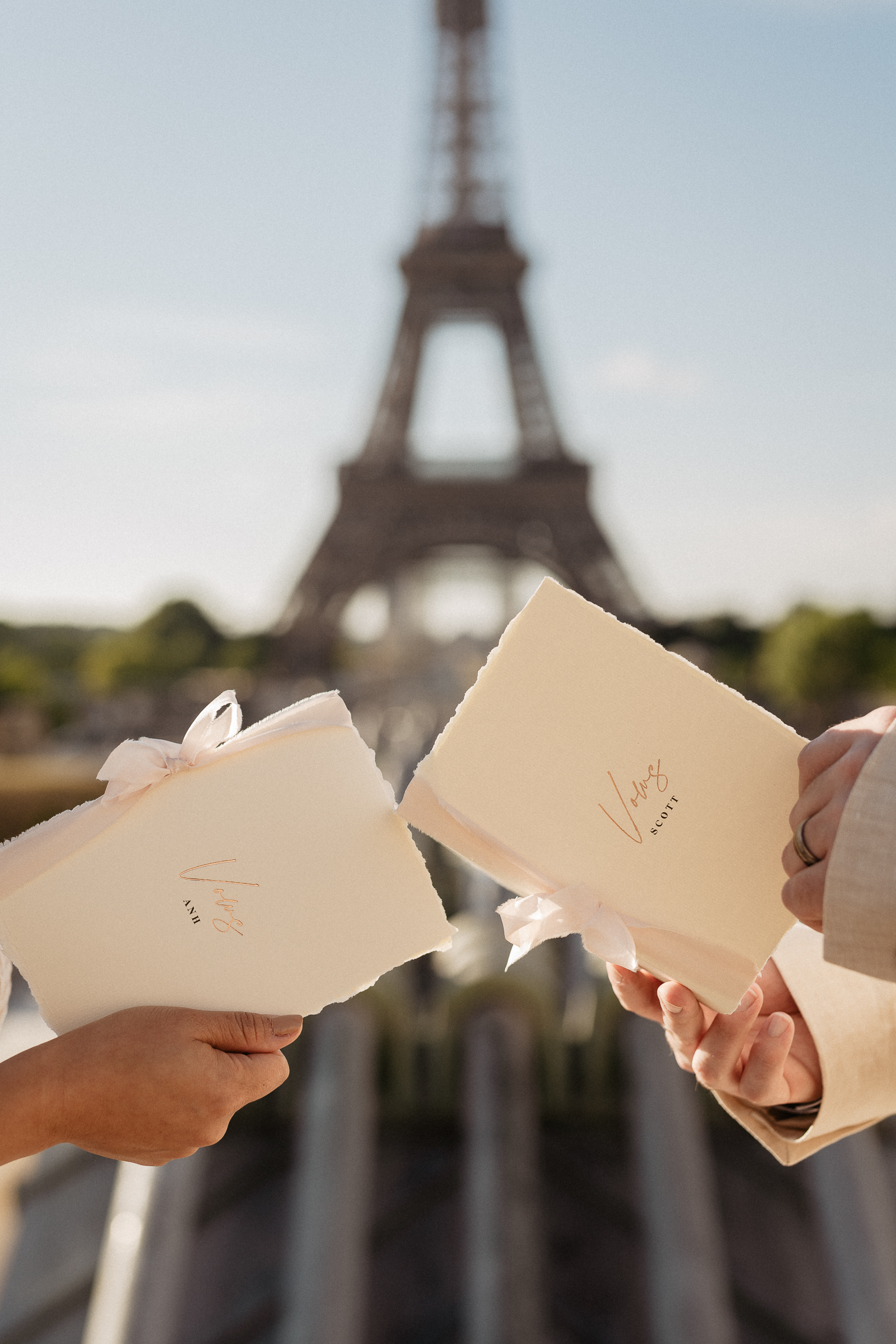Couple’s hands hold cream wedding invitations with the Eiffel Tower softly blurred behind in warm light