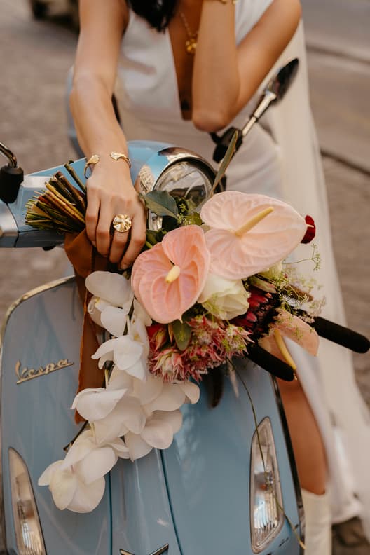 Bride in white leans on a blue Vespa on a Paris street, with pastel flowers and gold jewelry