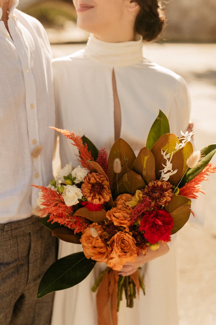 Bride in a cream dress holds a vivid autumn bouquet beside groom in warm daylight on a Paris street