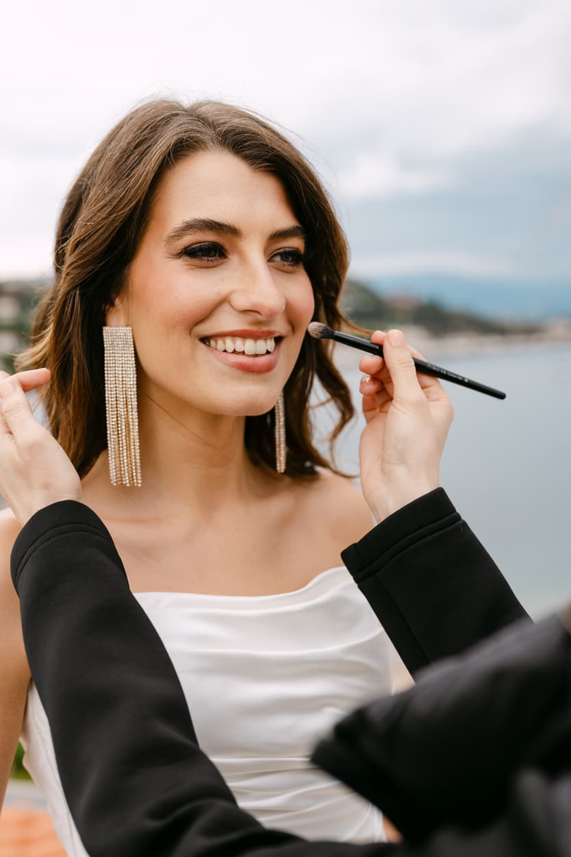 Bride in a white dress gets makeup touched up outdoors by a makeup artist near the water