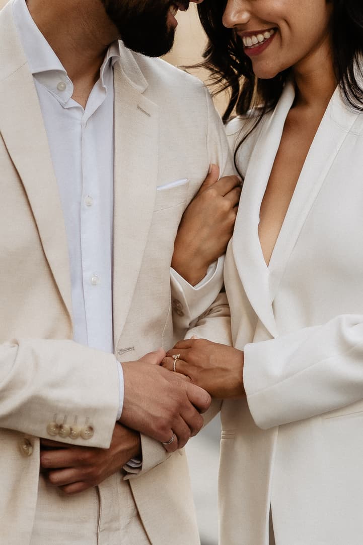 Bride and groom in cream suits hold hands in soft light, showing a wedding ring