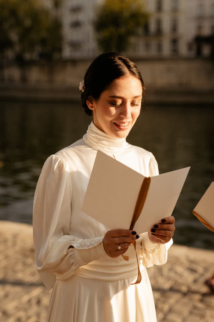 A bride in a white dress reads vows from a card beside the water in warm evening light
