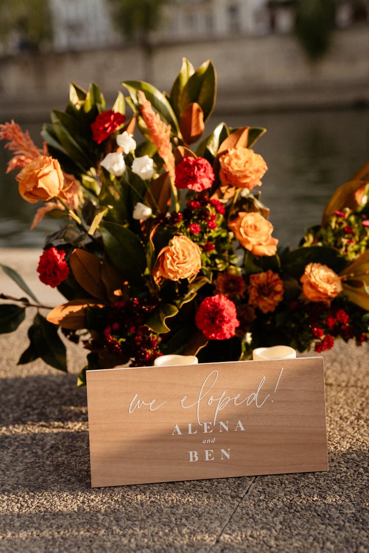 Sunlit floral arrangement beside a wooden sign reading “We eloped!” on a riverside quay
