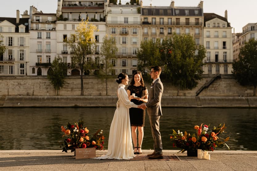 Bride and groom exchange vows with an officiant by the Seine, warm light on flower arrangements