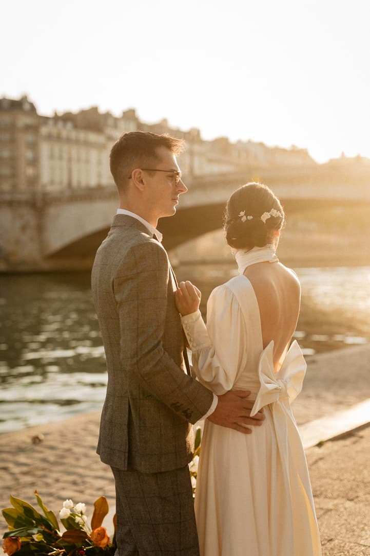 Bride and groom stand by the Seine at sunset, his hand at her waist and a bouquet on the stone path
