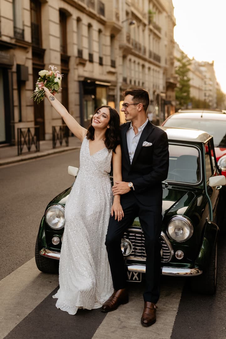 Bride and groom pose beside a vintage green car on a Paris street, her bouquet lifted in warm light