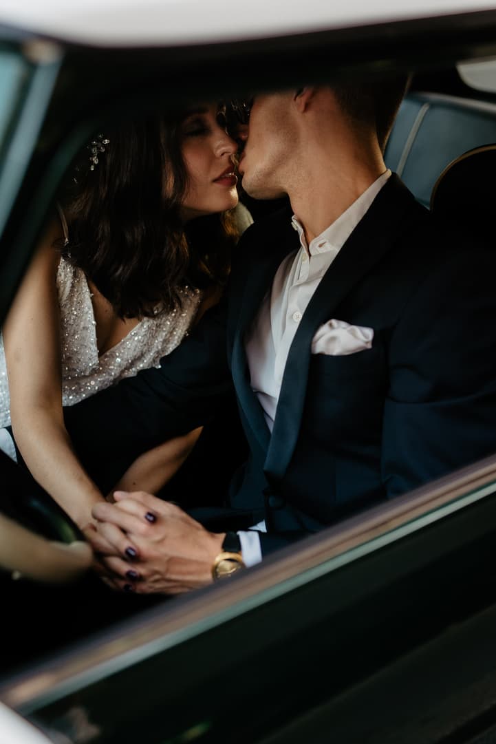 Bride and groom kiss in a dim car, her beaded dress and his suit framed by the window