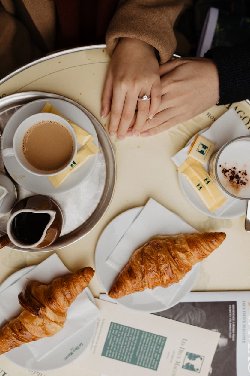 A couple holds hands over coffee and croissants at a café table, with an engagement ring visible