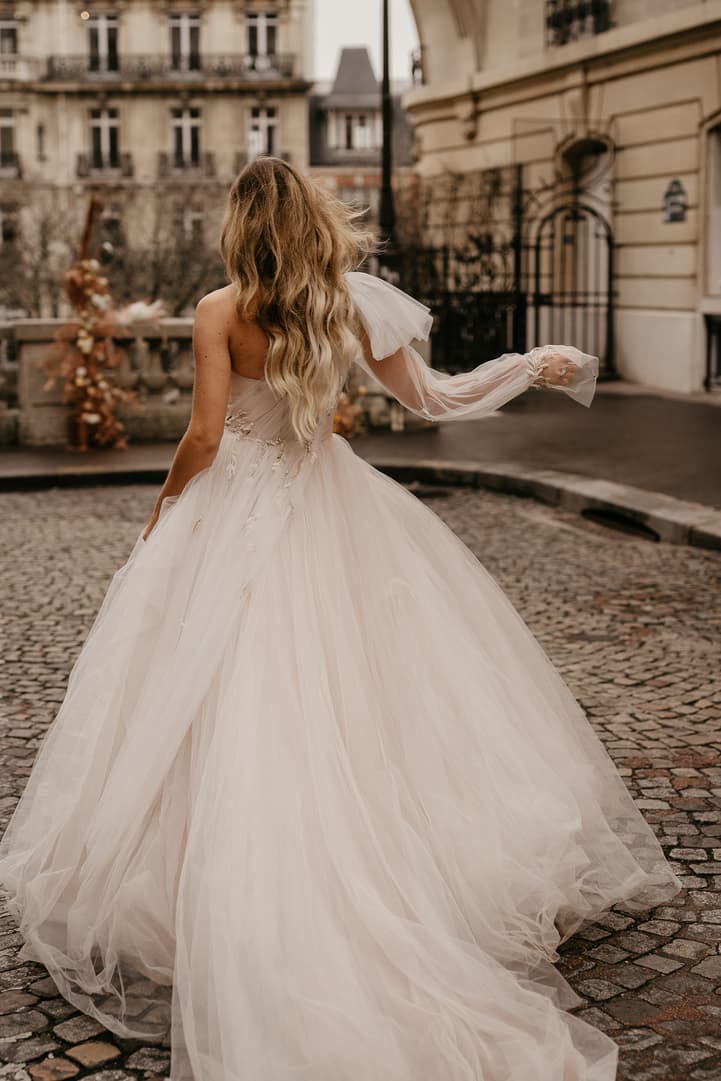 Bride in a flowing gown walks across a cobblestone Paris street in soft light, her veil trailing behind