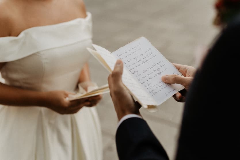 Bride and groom exchange handwritten vows on a Paris street in soft daylight, close on their hands