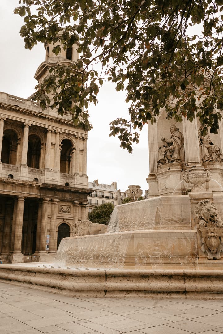 Stone fountain beneath leafy branches in a Paris square, with soft daylight and water spilling over tiers