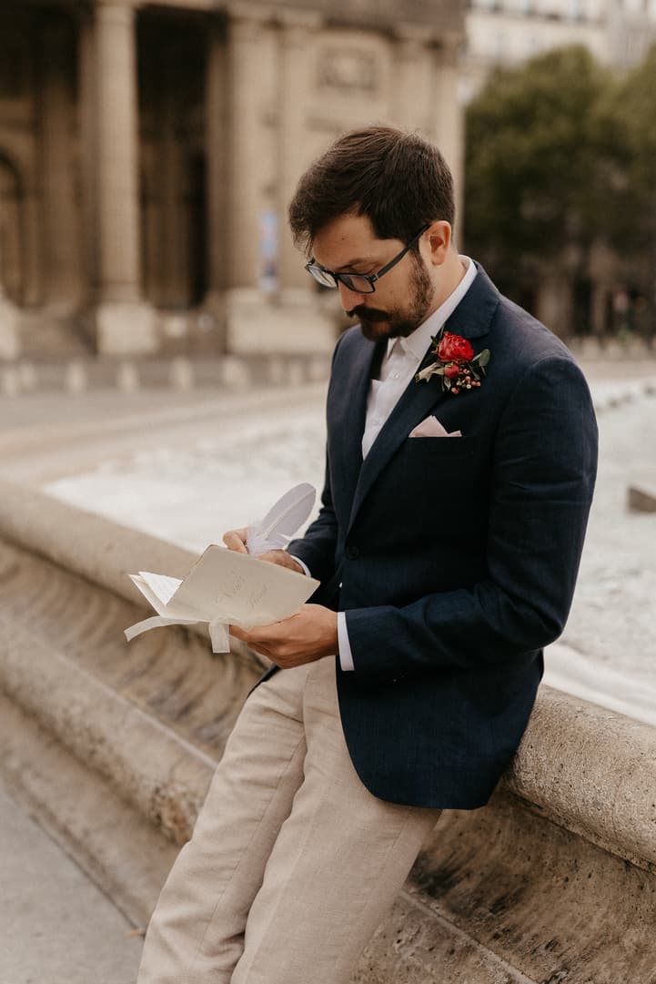 Groom in a navy suit reads a handwritten note on a Paris street in soft daylight, red boutonniere visible