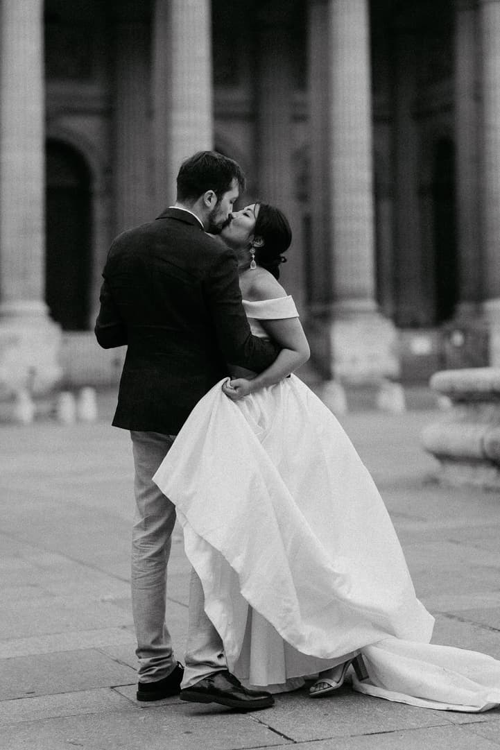 Black-and-white couple kissing on a Paris street, bride lifting her gown before stone columns