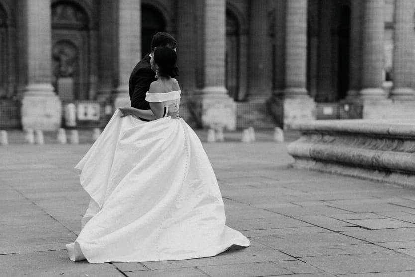 Bride and groom embrace on a Paris plaza, her flowing gown trailing across the stone ground