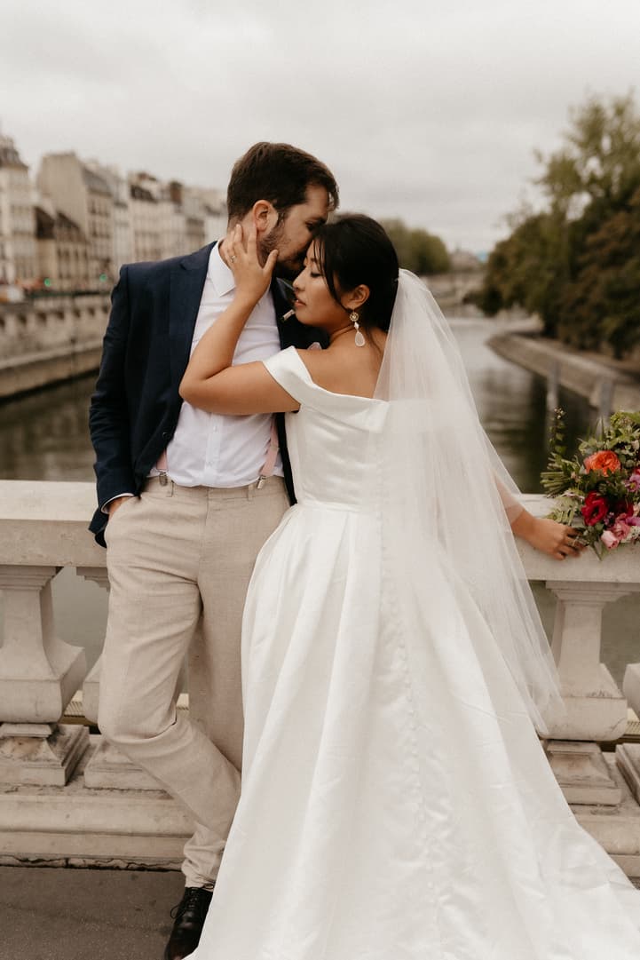 Bride and groom embrace on a Seine bridge in soft light, bouquet resting on the stone rail
