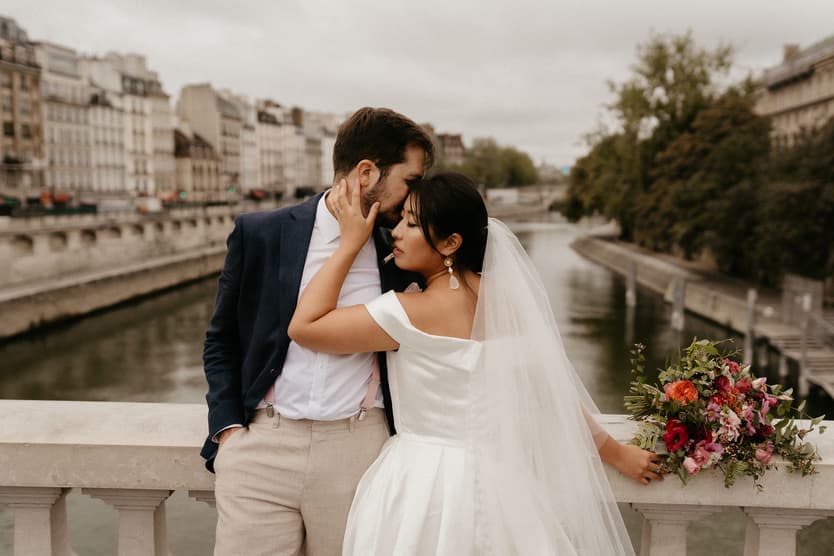 Bride and groom embrace on a bridge over the Seine, with a colorful bouquet and city buildings behind
