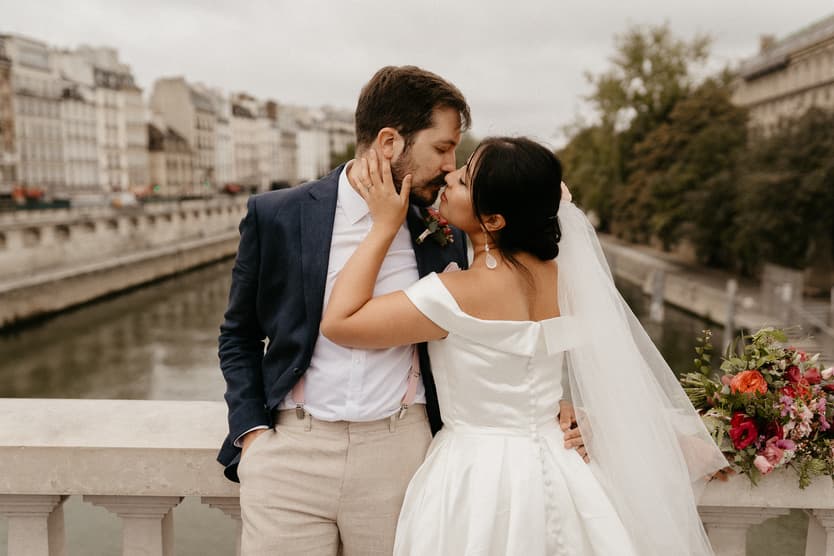 Bride and groom kiss on a bridge over the Seine, with soft overcast light and a bouquet nearby