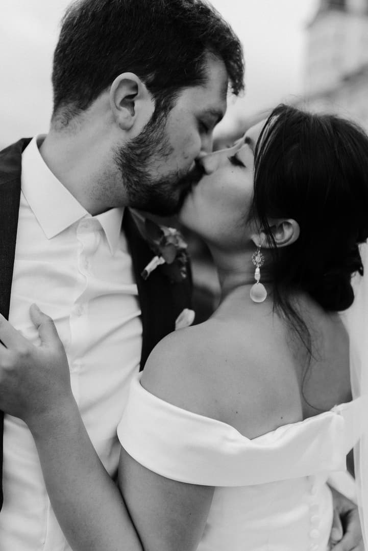 Bride and groom kiss in soft light on a Paris street, her off-the-shoulder dress and earrings visible