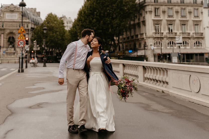 A bride and groom kiss on a Paris bridge, with the bride holding a bouquet in soft daylight