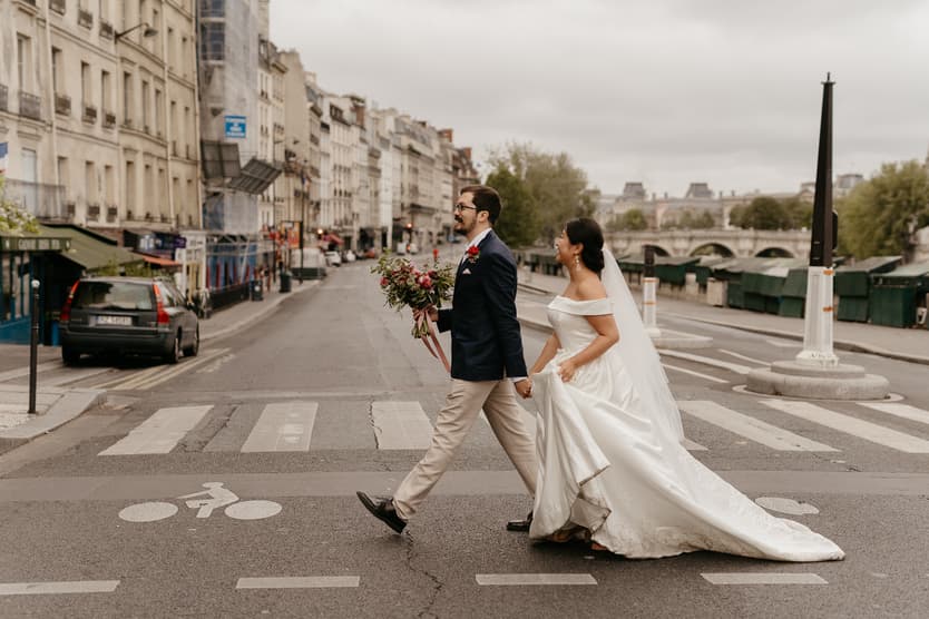 A bride and groom cross a Paris street beside the Seine under soft gray light