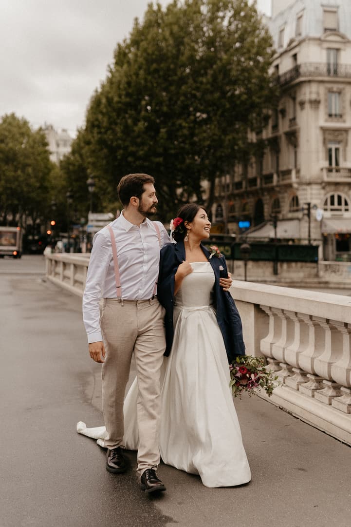 Bride and groom walk along a Paris bridge on an overcast day, bouquet in hand and pink suspenders showing