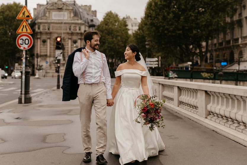Bride and groom walk hand in hand on a Paris street by the river, in soft light, bouquet in hand