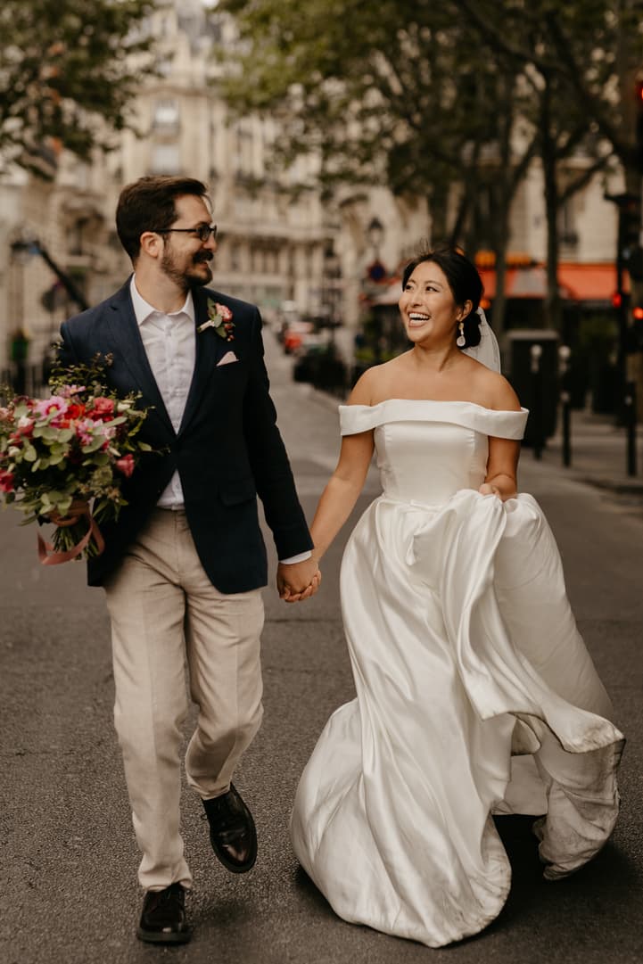 Bride and groom walk hand in hand down a Paris street, with the groom carrying a bouquet in warm light
