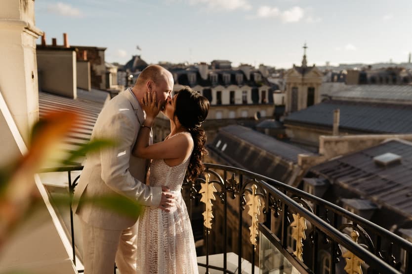 Bride and groom kiss on a Paris rooftop balcony in warm sunset light beside ornate iron railing