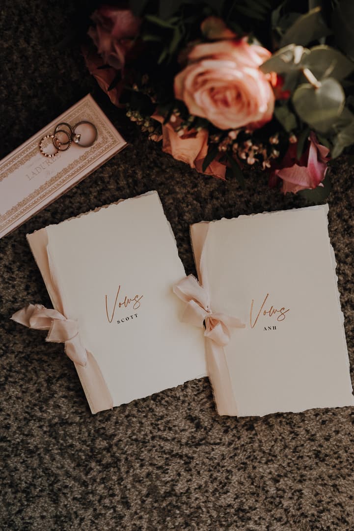 Two vow books tied with blush ribbon rest beside rings and a pink bouquet on a dark carpet