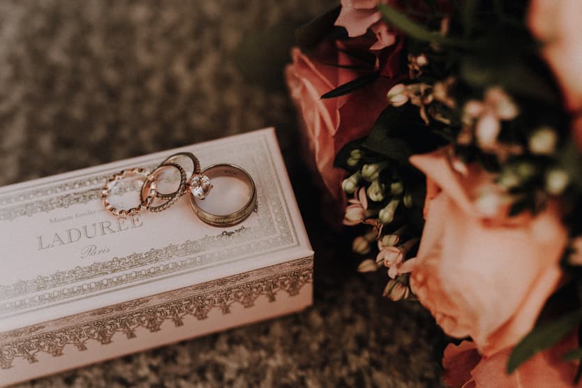 Wedding rings rest on a Ladurée box beside a peach bouquet in warm soft light