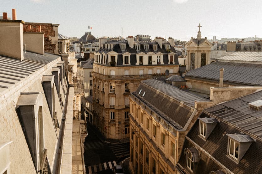 Sunlit Paris rooftops line a narrow street, with mansard windows and a small spire beyond