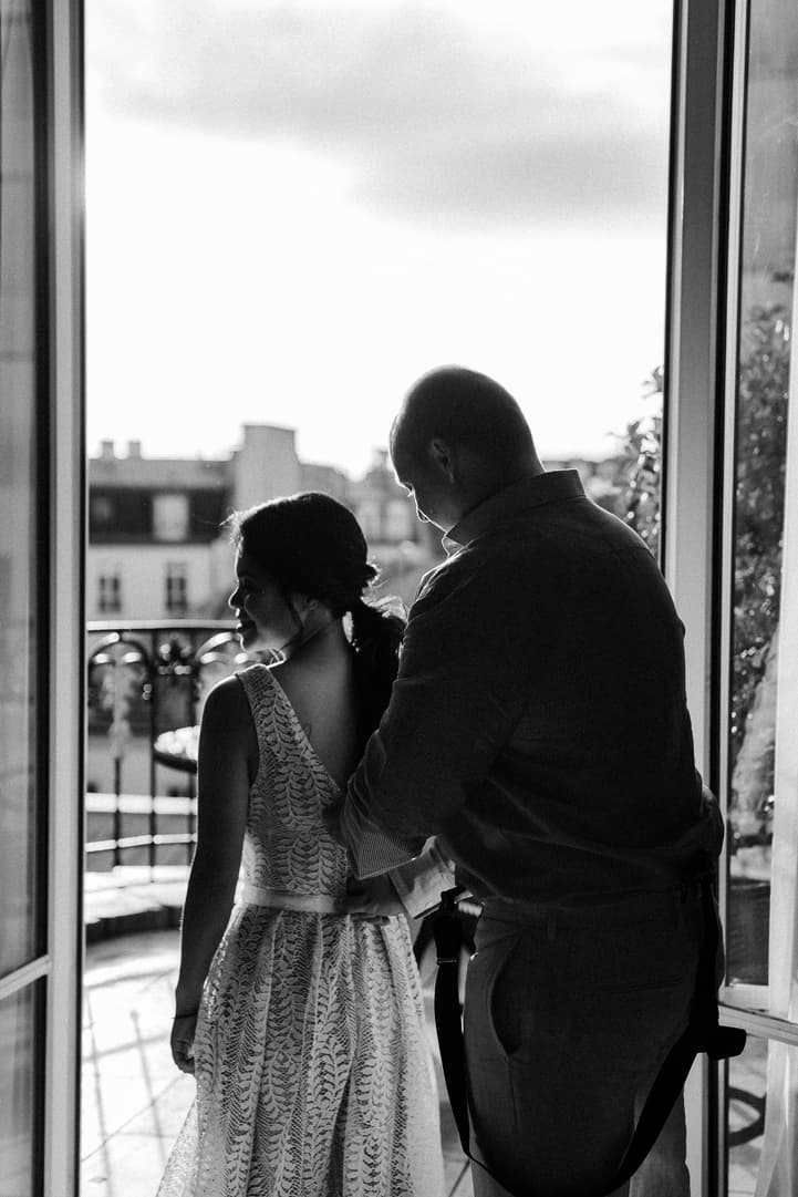 Bride and groom stand in a sunlit doorway, adjusting her dress on a Paris balcony overlooking rooftops