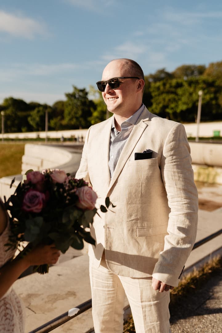 Groom in a light suit and sunglasses receives a bouquet in warm sunlight on a Paris terrace
