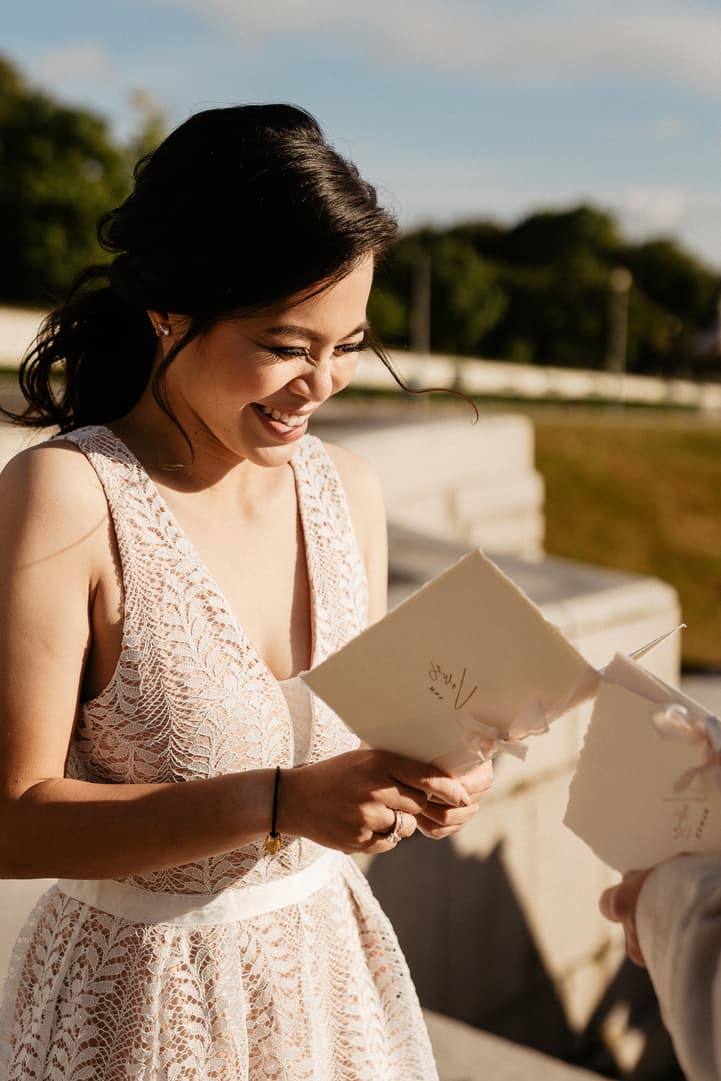 Bride in a lace dress smiles while reading a card outdoors in warm late-afternoon light on a Paris terrace
