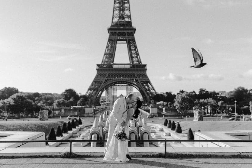 A bride and groom kiss in front of the Eiffel Tower, with a bird crossing a bright Paris sky