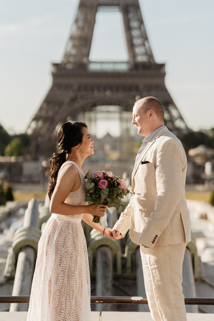 Bride and groom hold hands by the Eiffel Tower in warm light, bouquet of pink roses between them