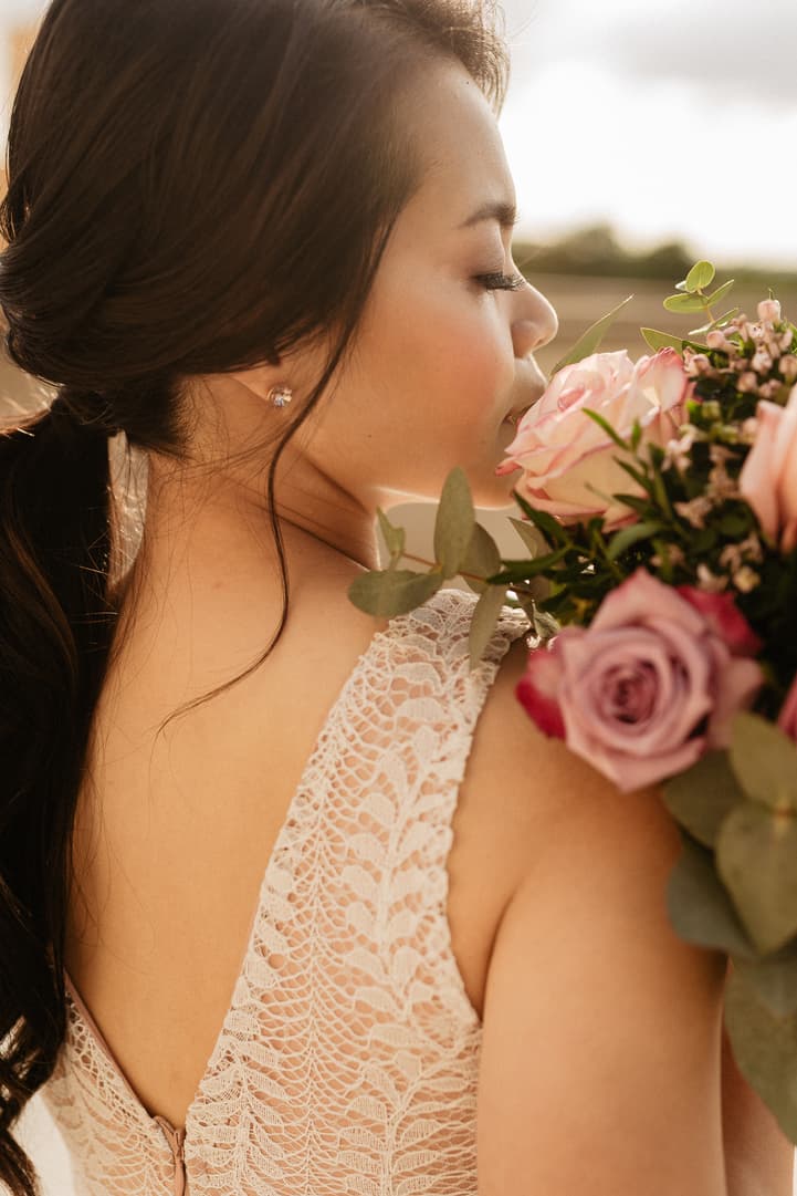 Bride in a lace dress smells a pink bouquet in warm golden light on a Paris terrace