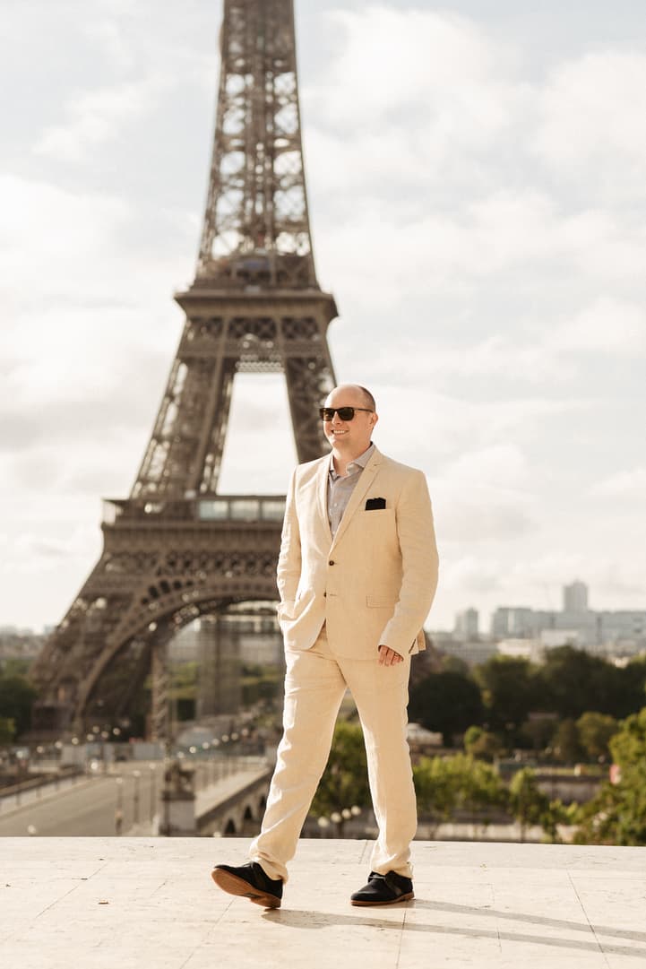 Groom in a cream suit and sunglasses stands before the Eiffel Tower in warm daylight