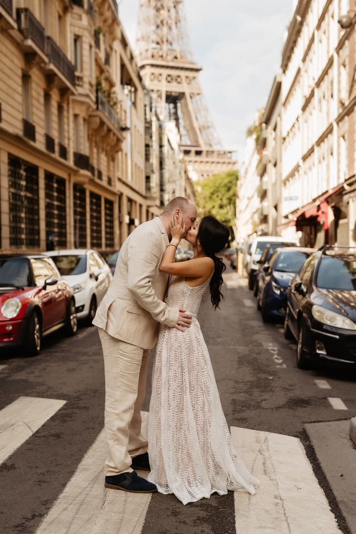 Bride and groom kiss on a Paris street with the Eiffel Tower rising behind parked cars
