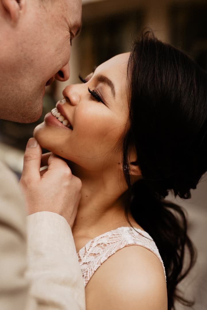Close-up of a couple embracing indoors, warm light on her lace dress and his hand at her chin
