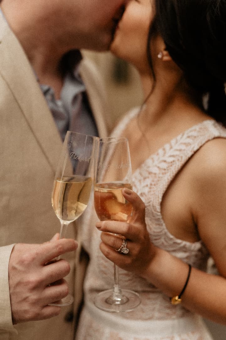 Bride and groom kiss while clinking champagne flutes, her ring catching warm indoor light