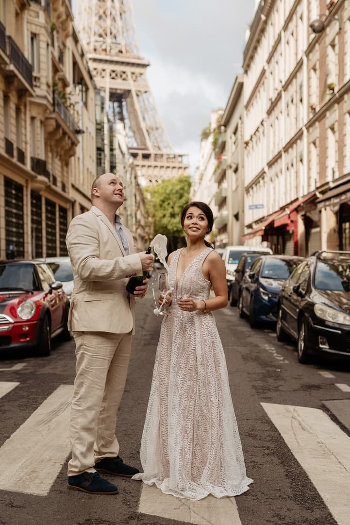 A couple toasts on a Paris street beneath the Eiffel Tower, with parked cars and soft daylight