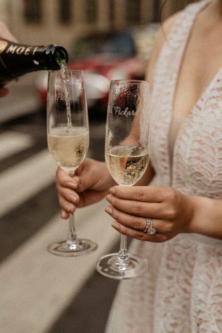 Bride in a lace dress holds two champagne flutes as sparkling wine is poured in warm city light