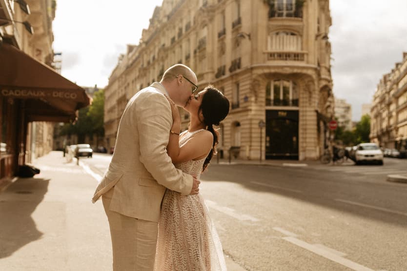 A couple in wedding attire kiss on a sunlit Paris street beside an ornate corner building
