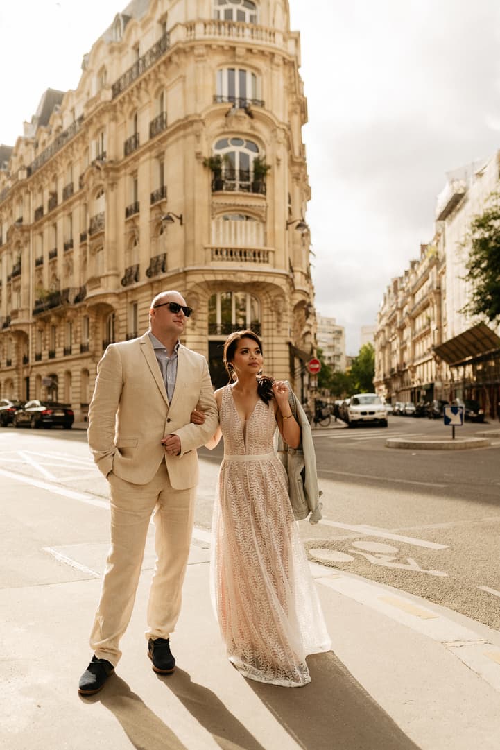 Bride and groom stand arm in arm on a Paris street, bathed in warm sunlight beside a grand corner building