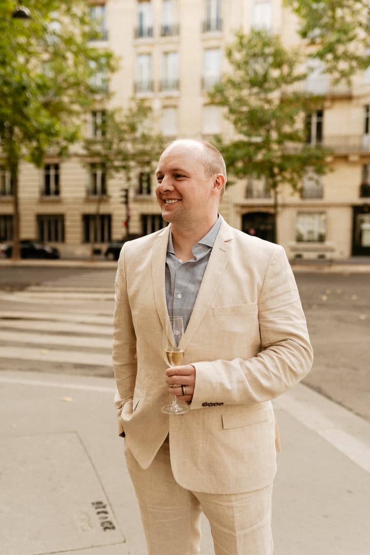 Groom in a beige suit holds a champagne flute on a soft-lit Paris street by a crosswalk
