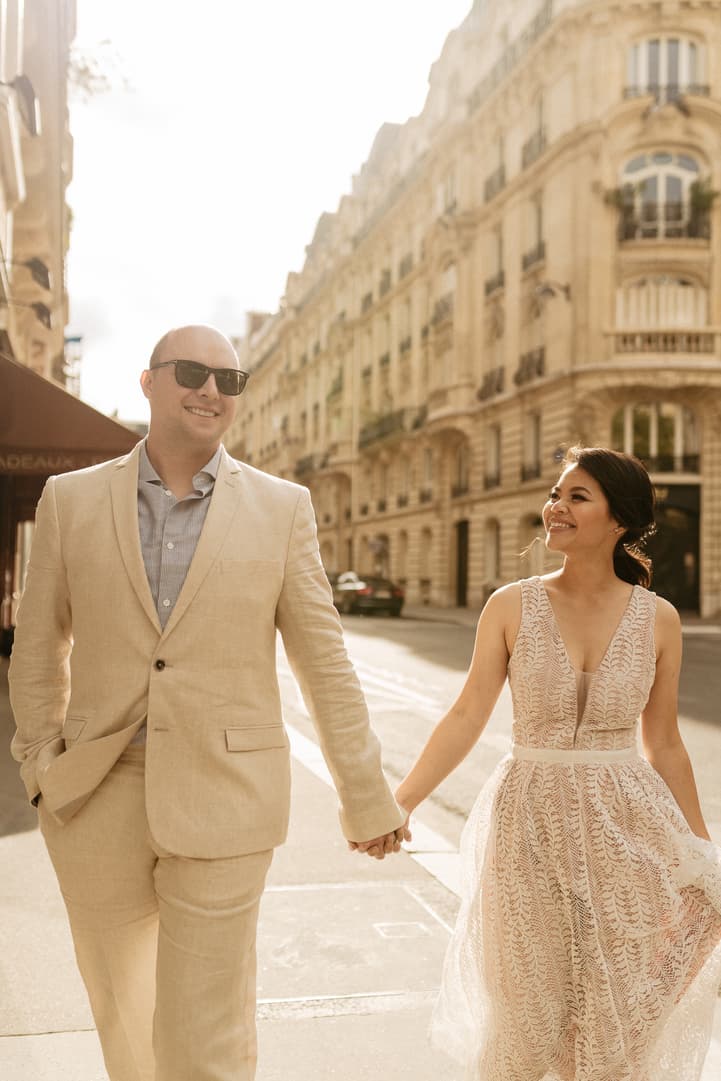 Bride and groom hold hands on a sunlit Paris street, smiling in beige wedding attire