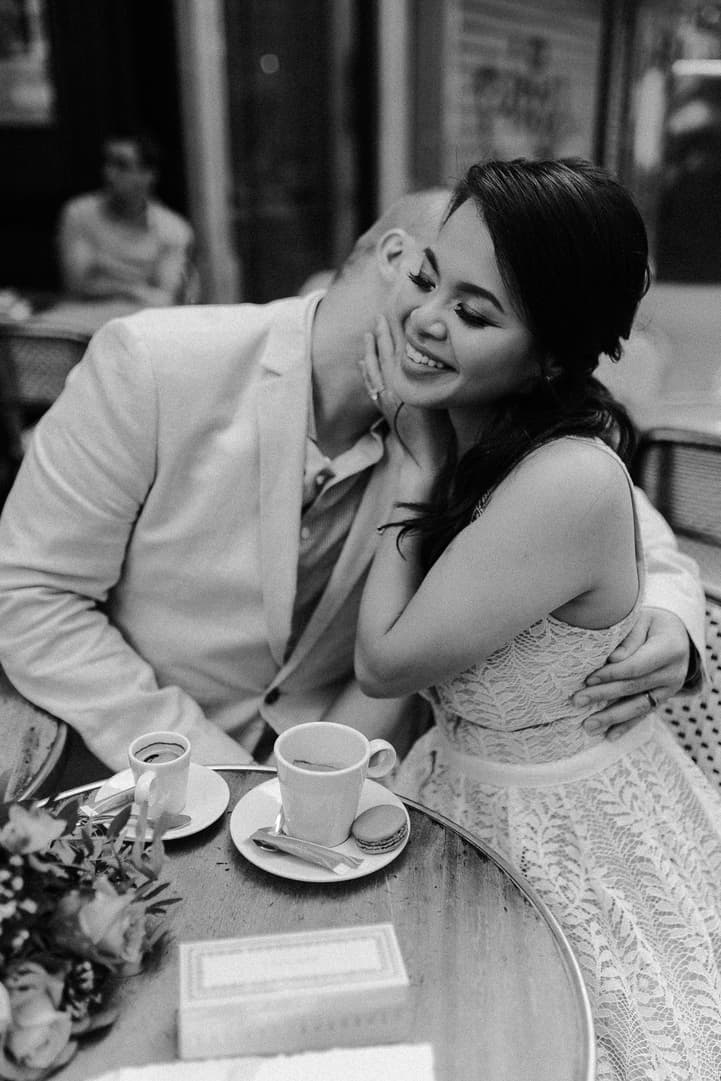 A bride and groom share a kiss at a Paris café table with coffee cups in soft light