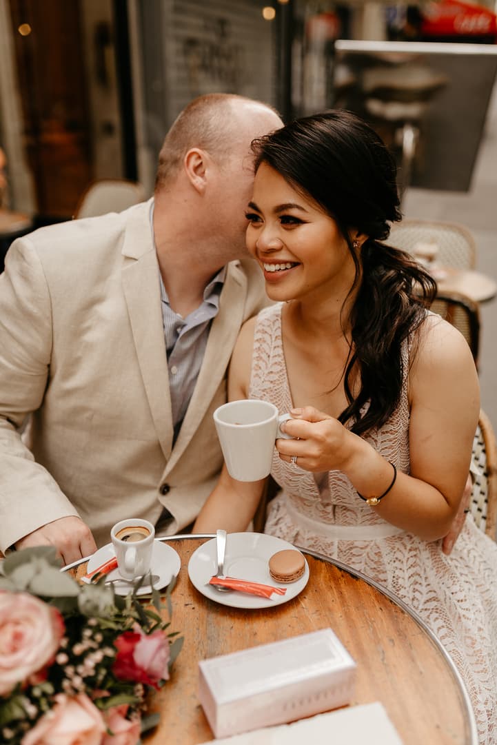Bride and groom sit at a café table, he leans in to whisper as she smiles with a cup in warm light