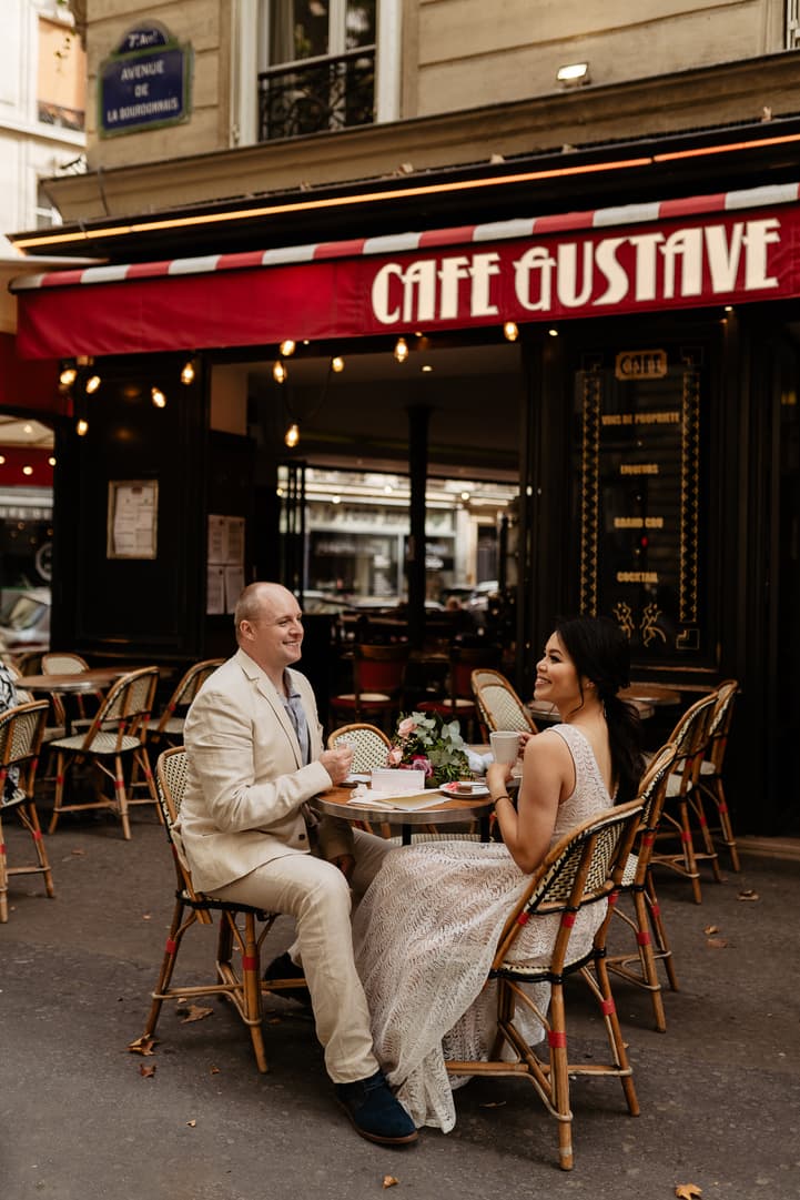 Bride and groom chat at a Paris café terrace under warm string lights, with flowers on their table